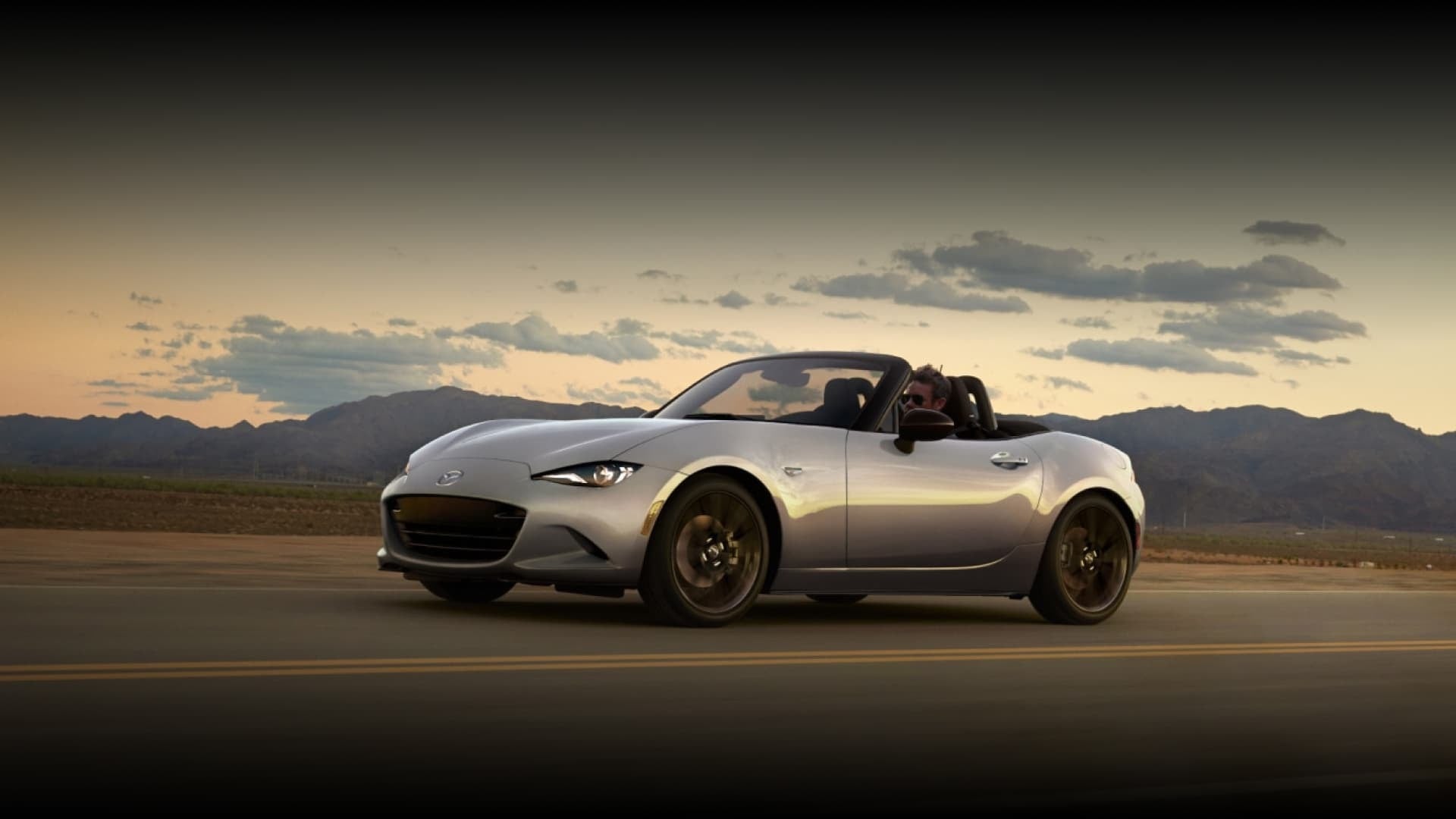 2025 Mazda MX-5 Miata, silver body, black soft-top, convertible, sleek design, sports car, roadster, open-road driving, scenic backdrop.